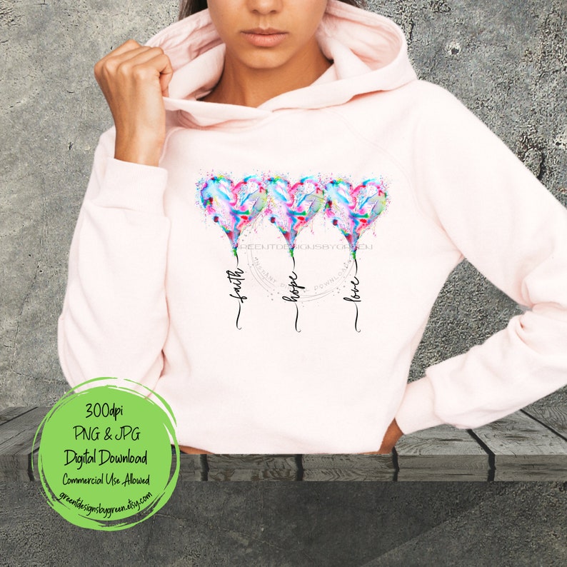 Pode incluir: Um moletom rosa com um design de tr&ecirc;s cora&ccedil;&otilde;es de aquarela com as palavras "f&eacute;", "esperan&ccedil;a" e "amor" escritas abaixo.