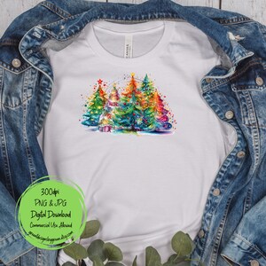 May include: A white t-shirt with a colorful watercolor design of three Christmas trees with lights and ornaments. The trees are surrounded by a white background with a few stars and snowflakes.