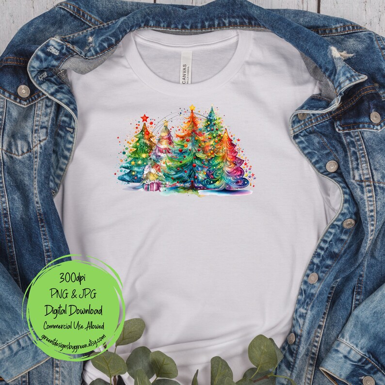 May include: A white t-shirt with a colorful watercolor design of three Christmas trees with lights and ornaments. The trees are surrounded by a white background with a few stars and snowflakes.