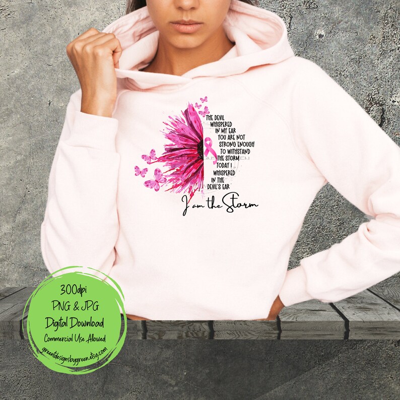 May include: Pink and white hoodie with a pink flower design and a pink ribbon in the center. The text "The devil whispered in my ear you are not strong enough to withstand the storm today I whispered in the devil's ear I am the storm" is printed on the hoodie.