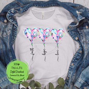 Pode incluir: Uma camiseta branca com um design de tr&ecirc;s bal&otilde;es de cora&ccedil;&atilde;o de aquarela com as palavras "f&eacute;", "esperan&ccedil;a" e "amor" escritas abaixo. Os bal&otilde;es s&atilde;o em tons de rosa, azul e verde.