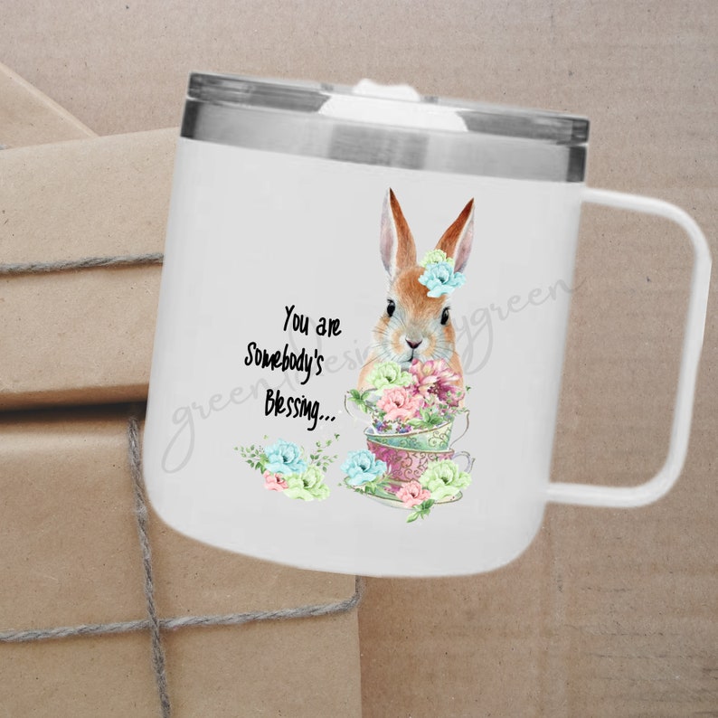 May include: White stainless steel travel mug with a handle. The mug features a watercolor illustration of a brown bunny rabbit holding a stack of teacups with flowers. The text "You are Somebody's Blessing..." is printed on the mug.