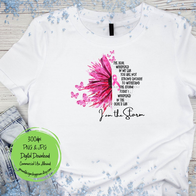 May include: A white t-shirt with a pink watercolor sunflower and a pink ribbon. The text "The devil whispered in my ear you are not strong enough to withstand the storm. Today I whispered in the devil's ear I am the storm" is printed on the shirt.
