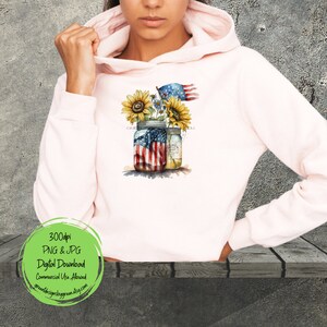 May include: A pink hoodie with a design of two mason jars with sunflowers and an American flag. The jars are decorated with the American flag. The text "God Bless America" is written on the hoodie.
