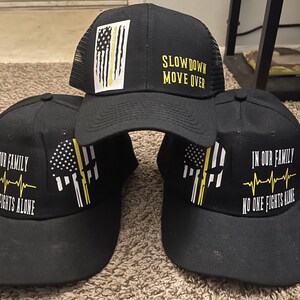 Puede incluir: Gorras de béisbol negras con varios diseños. Una gorra presenta el texto "SLOW DOWN MOVE OVER". Otras gorras muestran un diseño de calavera con una bandera estadounidense y el texto "IN OUR FAMILY NO ONE FIGHTS ALONE".