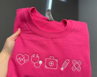 May include: A pink sweatshirt with white embroidered medical symbols including a heart, stethoscope, first aid kit, thermometer, and bandaid.