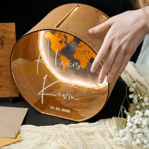 May include: A wooden wedding card box, cylindrical in shape, featuring a world map design and the names "Alexis & Kevin" with the date "10.03.2029". The interior is illuminated, creating a warm glow.