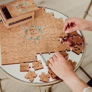 May include: A wooden puzzle with a floral design and the names Olivia & Ryan, along with the date 12.07.23. The puzzle pieces are arranged on a table and some are missing. The puzzle is a wedding guest book.