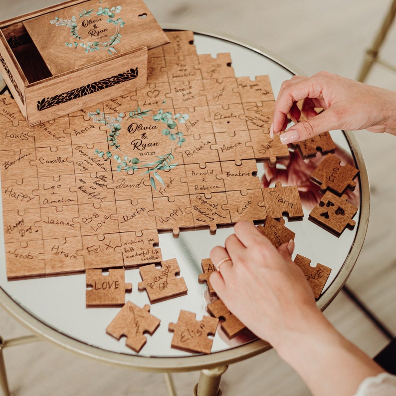 May include: A wooden puzzle with a floral design and the names Olivia & Ryan, along with the date 12.07.23. The puzzle pieces are arranged on a table and some are missing. The puzzle is a wedding guest book.