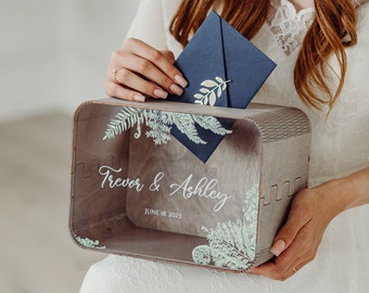 Può includere: Una scatola di legno per biglietti di matrimonio con un coperchio in acrilico trasparente. La scatola è decorata con un motivo a foglie e il testo "Trevor & Ashley" e "June 18, 2023". Una persona sta inserendo una busta blu nella scatola.