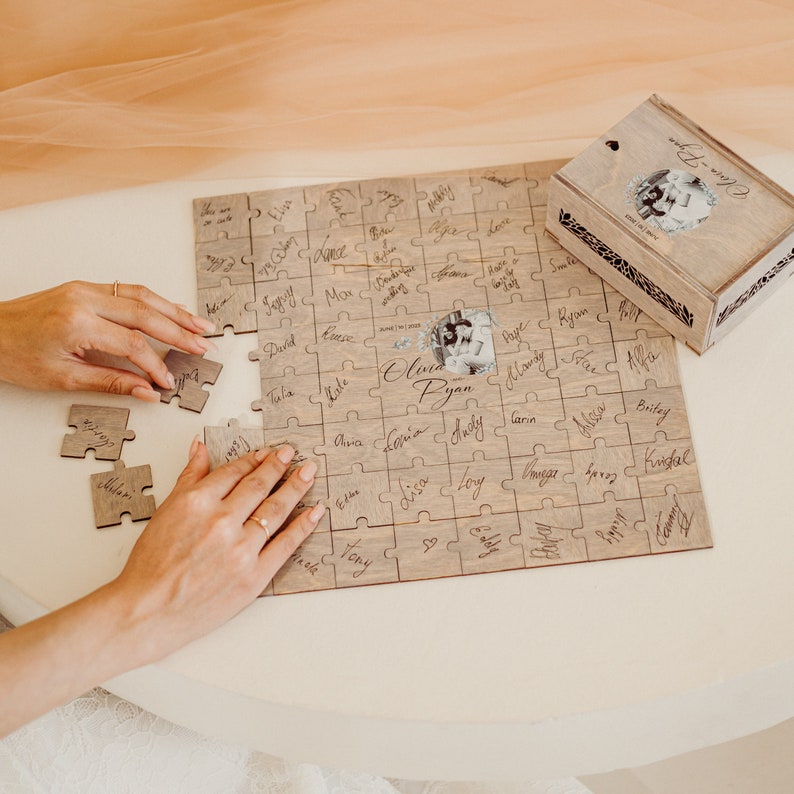 May include: A wooden jigsaw puzzle with a photo of a couple in the center. The puzzle pieces are signed with names. The puzzle is in a wooden box with a photo of the couple on the lid. The box is labeled "Olivia & Ryan".