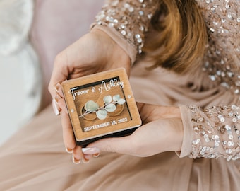 May include: A wooden ring box with a clear lid, featuring the names "Trevor & Ashley" and the date "SEPTEMBER 18, 2023". Two gold wedding bands are inside the box, with a sprig of eucalyptus leaves.