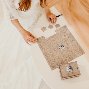 May include: A bride in a white lace dress places a puzzle piece on a wooden jigsaw puzzle with a photo of a couple and the words "Our Journey" written on it. The puzzle is on a white table with a small wooden box with a photo of a couple and the words "Our Journey" written on it.