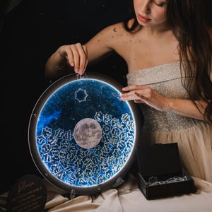 May include: A round, illuminated, galaxy-themed guest book with a moon and stars design. The guest book is made of clear acrylic and has a black frame. The moon features the names "Alisa & Celeste" and the date "17.11.24".
