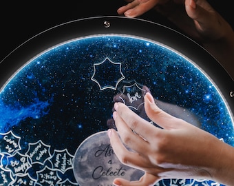 May include: A circular, illuminated art piece with a starry night design. The piece features a blue and white galaxy background, with a moon and the names "Asa & Celeste" and the date "12/12" inscribed. The word "love" is written on a star-shaped piece.