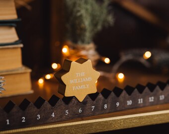 Op de afbeelding: Een gouden stervormige ornament met de tekst "The Williams Family" erop. De ornament staat op een houten plank met een nummerenschaal.