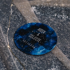 Könnte beinhalten: Ein rundes, blaues und weißes Holzschild mit einem Galaxie-Design. Das Schild trägt die Aufschrift "Aster & Celeste 12 | 12 | 24" in weißen Buchstaben.