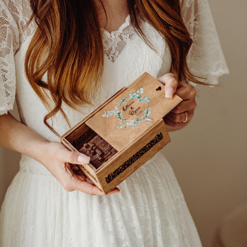 May include: A wooden box with a floral design and the names Olivia & Ryan engraved on the lid. The box is open and has a heart-shaped cutout on the lid. The box is being held by a person wearing a white dress.
