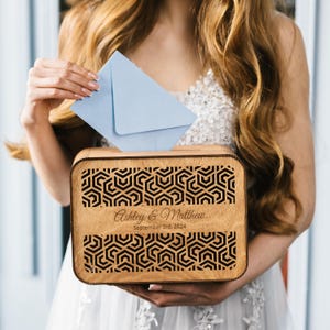 Könnte beinhalten: Eine hölzerne Hochzeitskartenbox mit geometrischem Muster und den eingravierten Namen "Ashley & Matthew" sowie dem Datum "September 3rd, 2024". Ein hellblauer Umschlag wird in die Box gelegt.