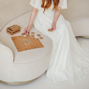 May include: A woman in a white lace dress sits on a cream-colored couch and assembles a wooden jigsaw puzzle with a floral design. The puzzle is on a light brown surface. A small wooden box with a floral design is on the couch next to the puzzle.