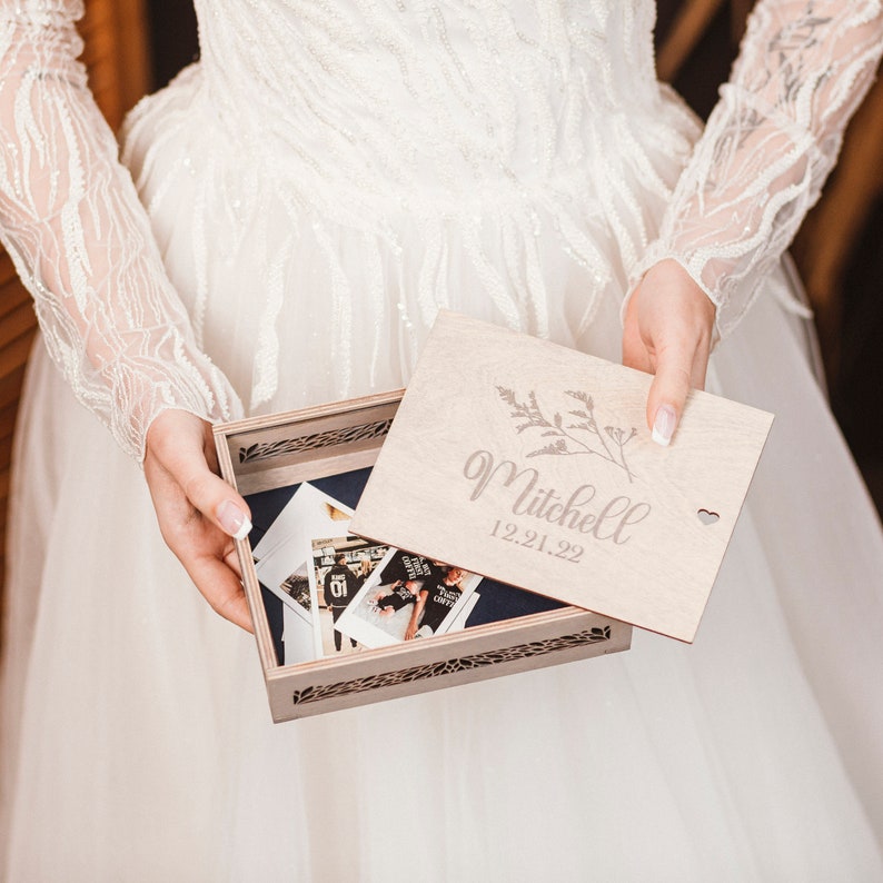 May include: A bride holds a wooden keepsake box with a lid that reads "Mitchell 12.21.22". The box is decorated with a floral design and has a small heart cutout. The box is open and contains several photographs.