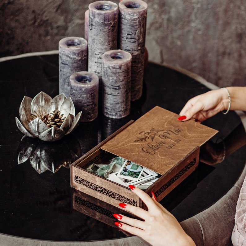 May include: A wooden photo box with a lid that has the words "Our Love" and the date "12.12.12" engraved on it. The box is open and there are photos inside. The box is sitting on a black table with candles in the background.