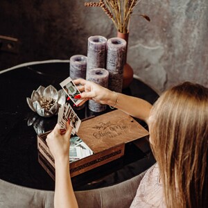 May include: A wooden photo box with a personalized inscription "Olivia Mae 12.21.22" and a floral design. The box is open and filled with instant photos.
