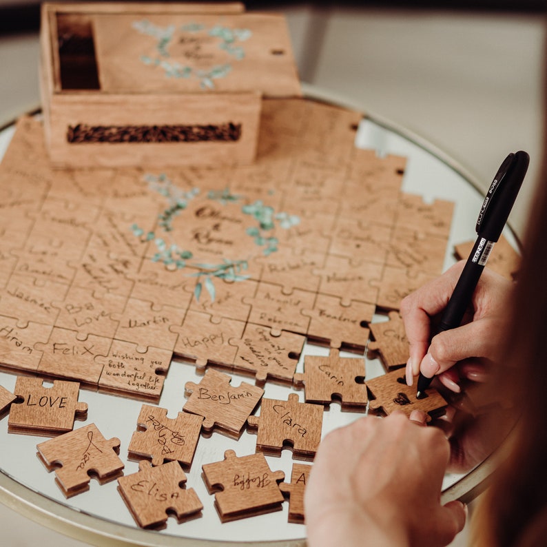 May include: A wooden puzzle with individual pieces that guests can sign at a wedding. The puzzle is assembled on a table with a wooden box and a black pen.