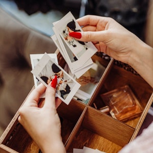 May include: A wooden box with multiple compartments filled with instant photos. The photos are of a couple and a baby. The box is open and a hand is reaching in to grab a photo.