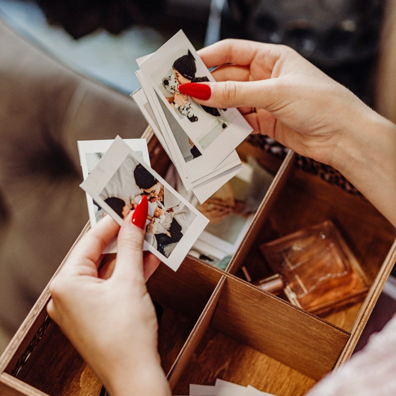 May include: A wooden box with multiple compartments filled with instant photos. The photos are of a couple and a baby. The box is open and a hand is reaching in to grab a photo.