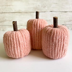May include: Three decorative pumpkins crafted from soft, light pink ribbed fabric. Each pumpkin features a distinct, dark brown natural twig stem protruding from the center top. The pumpkins are arranged in a cluster, showcasing a textured surface.