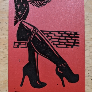 Pode incluir: Uma impressão de linóleo em preto e branco de uma mulher usando sapatos de salto alto e um chapéu estampado. O fundo é vermelho com um padrão de tijolo preto.