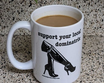 May include: White ceramic mug filled with coffee. The mug features the text "support your local dominatrix" in black, along with a graphic of high-heeled boots. The website address maenadpress.etsy.com is printed below the graphic.