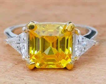 May include: A yellow gemstone engagement ring with a square emerald cut center stone and two triangular side stones. The ring is set in a silver band with gold accents.