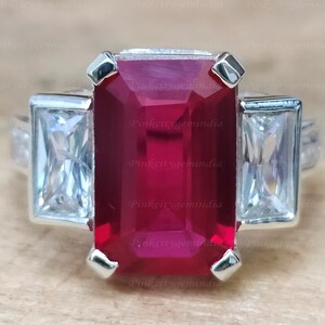 Peut inclure: Une bague en argent avec une grande pierre de rubis taillée en émeraude entourée de deux diamants rectangulaires plus petits.
