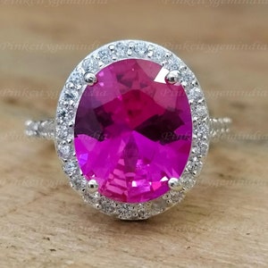 Pode incluir: Um anel de prata com uma grande gema oval rosa cercada por um halo de gemas brancas menores.