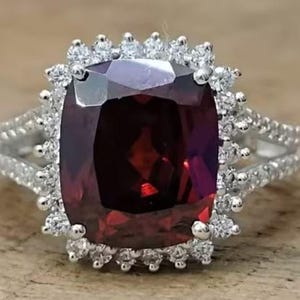 May include: A silver ring featuring a large, dark red gemstone encircled by a halo of smaller, clear gemstones.