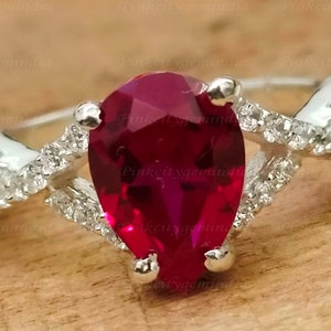 Peut inclure: Une bague en argent avec une pierre rouge en forme de poire entourée de petites pierres blanches. La bague a une bande torsadée.