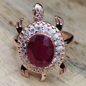 May include: A rose gold ring featuring a turtle design with a large red gemstone surrounded by smaller white gemstones.
