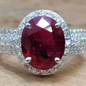May include: A silver ring with a large oval ruby gemstone surrounded by a halo of smaller white gemstones. The band of the ring is also set with smaller white gemstones.