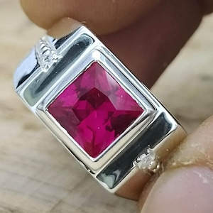 Puede incluir: Un anillo de plata con una piedra preciosa roja brillante de forma cuadrada engastada en el centro. El anillo tiene un diseño simple y elegante.