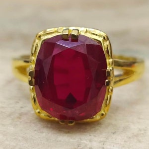 Peut inclure: Une bague en or avec une grande pierre précieuse rouge foncé sertie dans une bélière carrée. La bague a une bande simple.