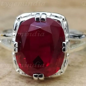 Può includere: Un anello d'argento con una grande pietra preziosa rosso scuro incastonata in una montatura quadrata. L'anello ha un design decorativo e ornato.
