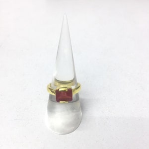 Może przedstawiać: Złoty pierścionek z czerwonym kamieniem szlachetnym w kwadratowym kształcie. Pierścionek jest prezentowany na przezroczystym plastikowym stojaku na pierścionki.