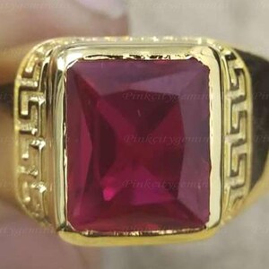 Könnte beinhalten: Ein goldfarbener Ring mit einem großen, quadratischen, tiefroten Edelstein. Der Ring hat einen dekorativen Rand mit einem sich wiederholenden Muster. Der Edelstein ist der Blickfang und bildet einen auffälligen Kontrast zur Goldfassung.