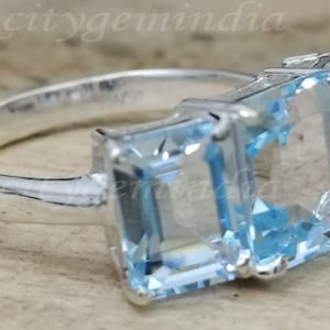 May include: A silver ring with three light blue gemstones. The gemstones are emerald cut and set in a prong setting.