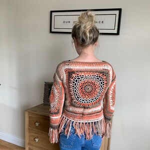 May include: A crocheted top with a brown, orange, and white color scheme. The top has a square design with a circular pattern in the center. The top has fringe along the bottom and sleeves.
