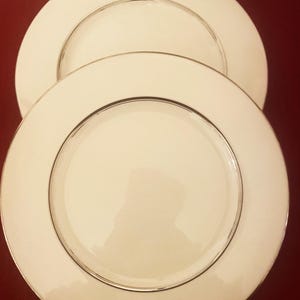 May include: Two ivory-colored ceramic plates with a silver-colored trim. The plates are round and have a simple, elegant design. The plates are stacked on top of each other, with the top plate slightly overlapping the bottom plate.