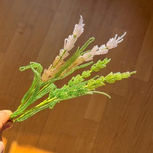 May include: A handcrafted beaded floral arrangement featuring green stems and leaves, with light pink and white beaded flower heads. The arrangement is held against a wooden background, with sunlight casting shadows.