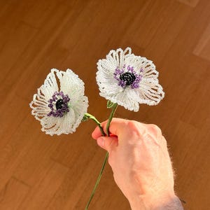 Könnte beinhalten: Zwei handgefertigte, weiße, perlenbesetzte Blumen mit lila und schwarzen Zentren werden vor einem braunen Holzhintergrund gehalten. Die Blumen haben zarte, detaillierte Blütenblätter und grüne Stiele. Ein einzigartiger Bastelartikel.